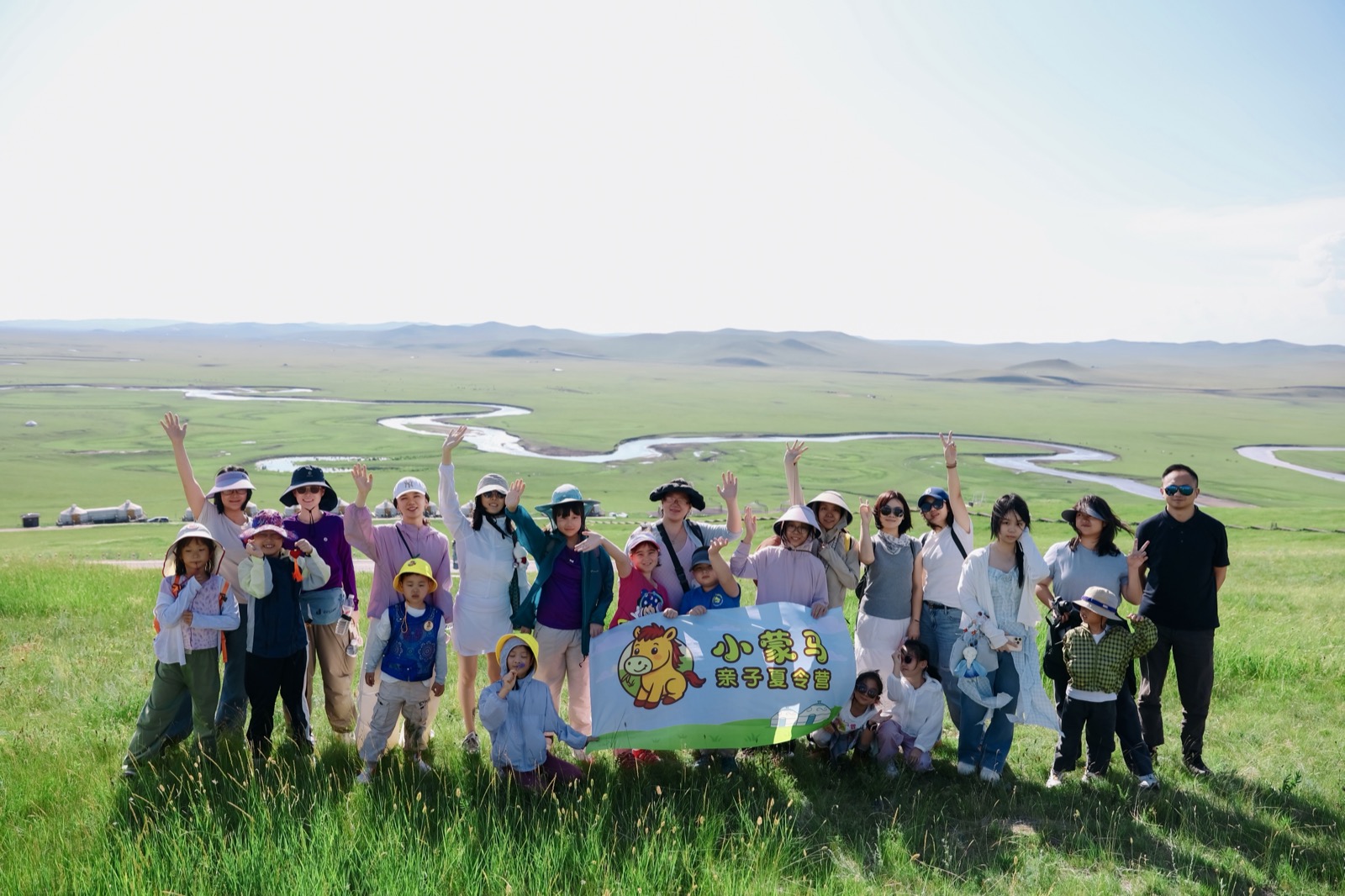 小蒙马亲子夏令营在莫日格勒河旁举旗合影，背景是蜿蜒的草原河流